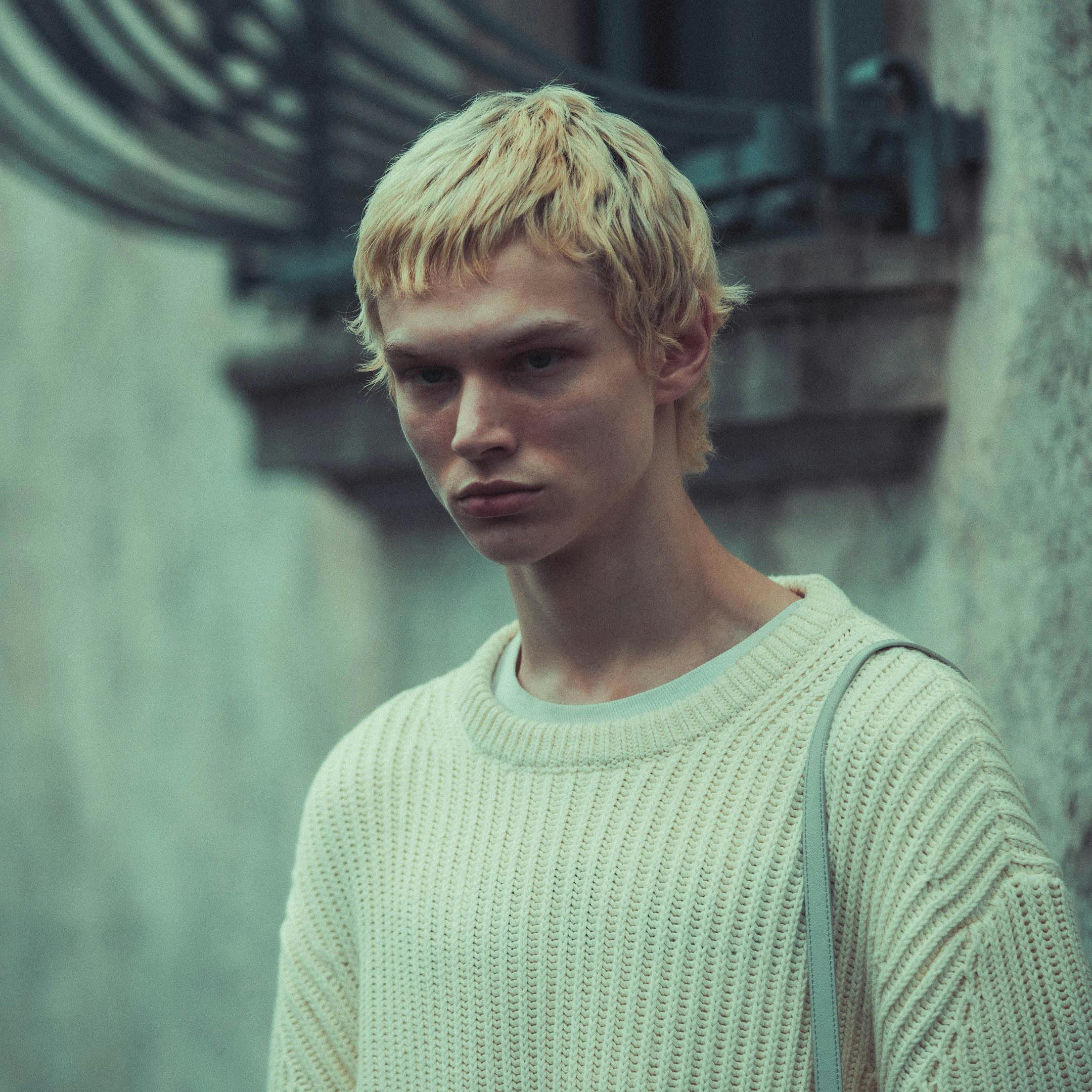 Close-up of a blonde man wearing a knit sweater outdoors with a moody background.
